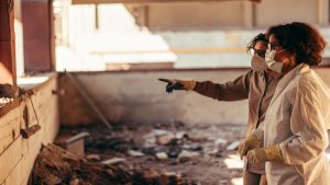 Two people wearing protective masks and gloves inspect a damaged indoor area, with one person pointing toward debris on the ground as they assess the cleanup needs.