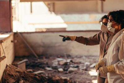 Two people wearing protective masks and gloves inspect a damaged indoor area, with one person pointing toward debris on the ground as they assess the cleanup needs.