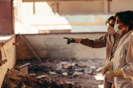 Two people wearing protective masks and gloves inspect a damaged indoor area, with one person pointing toward debris on the ground as they assess the cleanup needs.