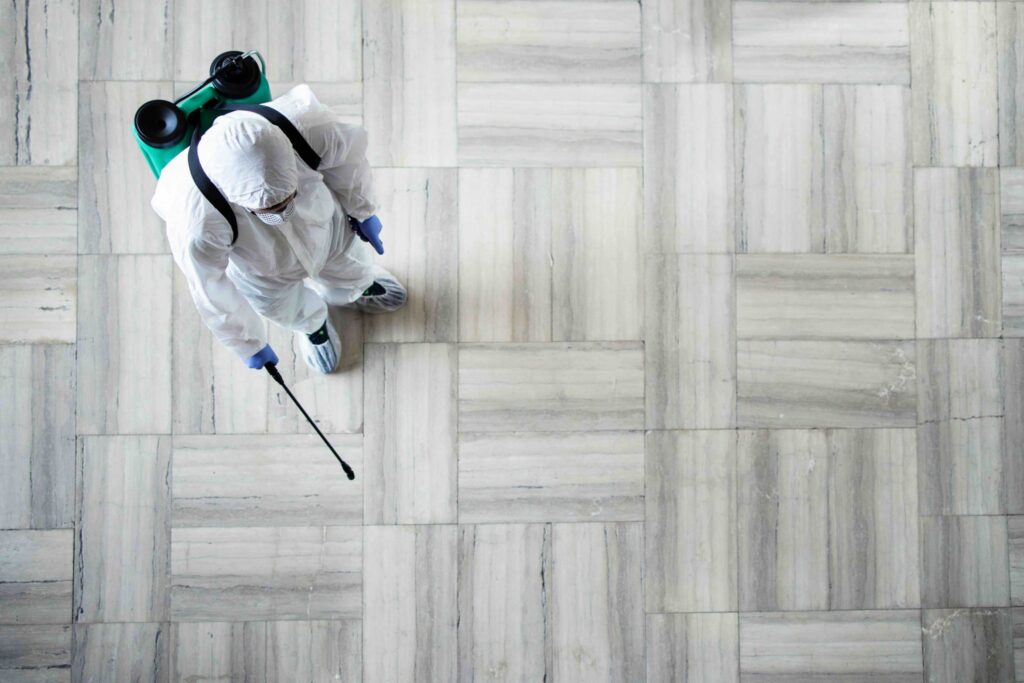 A person in protective gear sprays a surface with a pest control device on a tiled floor.