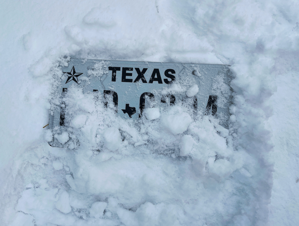 A Texas license plate partially buried in snow, with only a few letters visible.