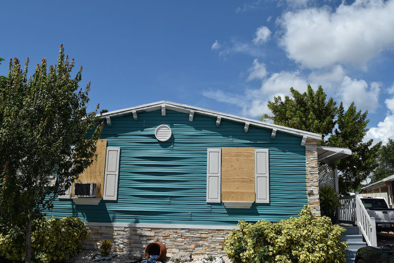 A teal house with boarded windows, surrounded by greenery, under a partly cloudy sky, prepared for a hurricane.
