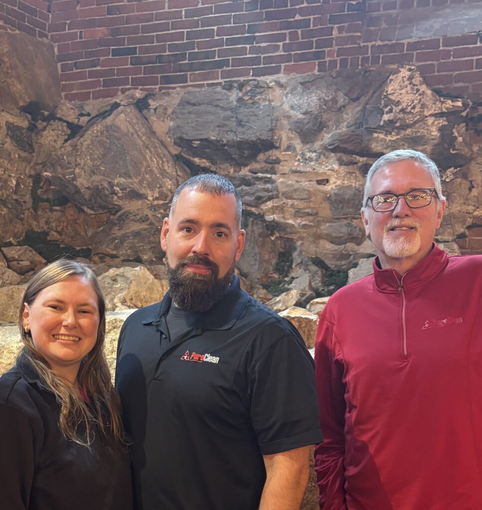 Three smiling PuroClean team members pose together in front of a rustic stone and brick wall.