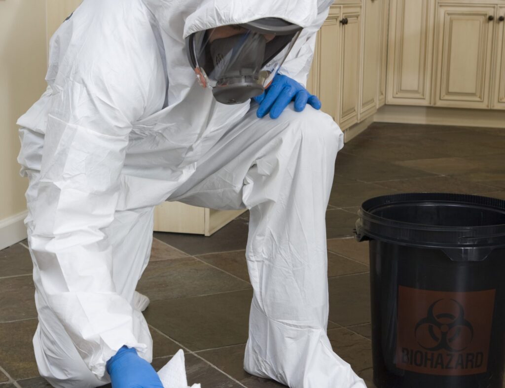 A person in a protective suit and mask kneels on a floor, cleaning with gloves and a bucket labeled with a biohazard symbol.