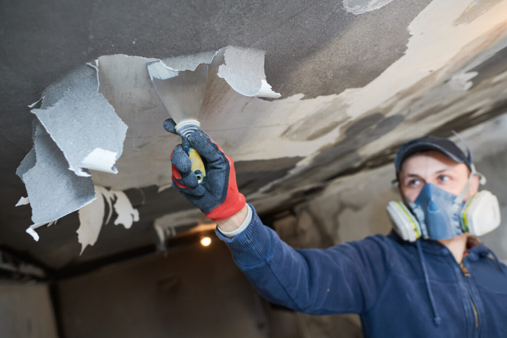 A worker wearing a mask and gloves removes peeling paint from a ceiling with a scraper.