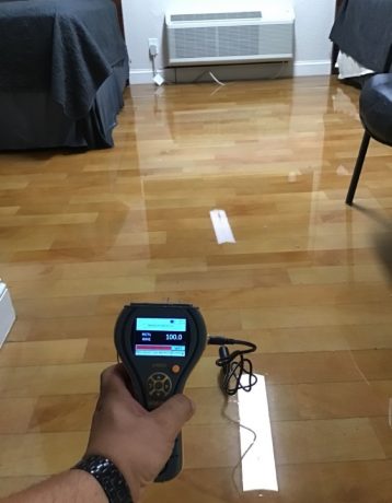 A moisture meter held over a wet wooden floor shows a 100% moisture reading.