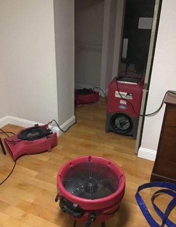 Red industrial fans and a dehumidifier dry a wooden floor after water damage. TITLE