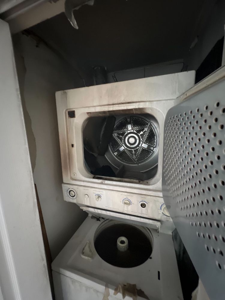 A smoke-damaged stacked washer and dryer unit with open doors in a dark utility closet