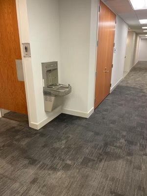 A silver water fountain is mounted on a white wall in a carpeted office hallway, near a