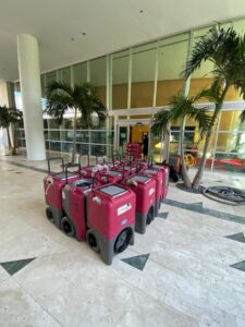 A row of red industrial dehumidifiers sits on a marble floor in a modern building lobby with palm trees.
