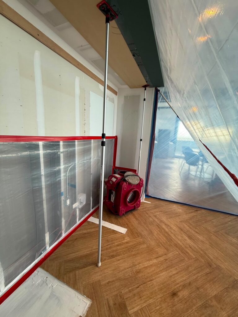 A room with temporary plastic dust barriers, a red air mover, and unfinished drywall.