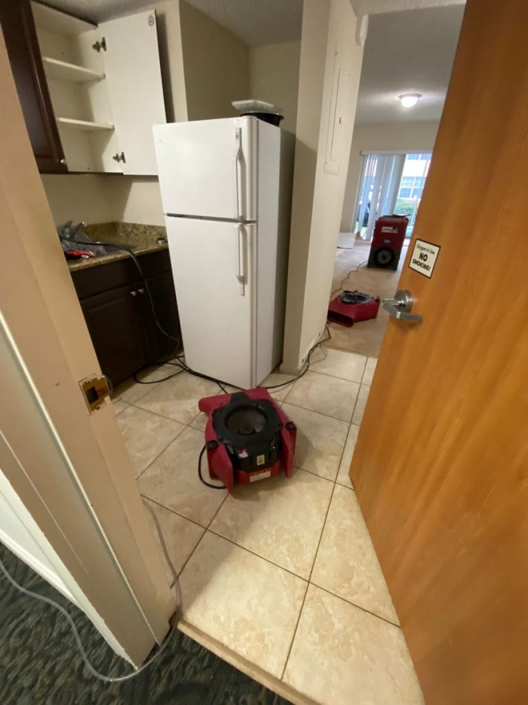 Red industrial drying fans are operating in a kitchen and adjacent room with a tiled floor. TITLE
