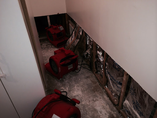 Red air movers are positioned in a water-damaged room with exposed walls and flooring.