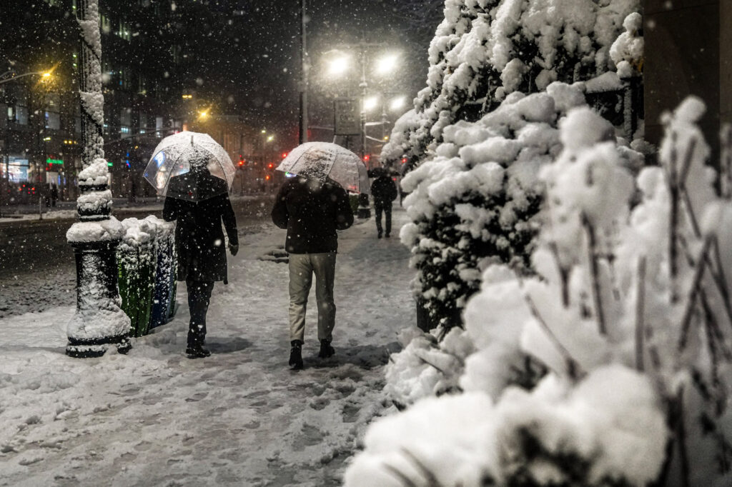 Two people walk on a snowy street at night, holding umbrellas amidst falling snow, with city lights in the background.