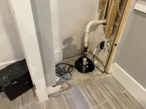 A sump pump basin with PVC pipe and electrical cords is installed in a partially exposed wall corner.