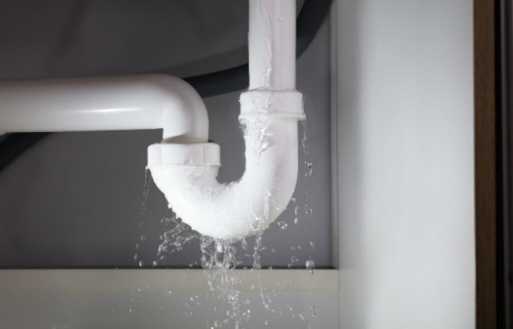 A white plastic plumbing pipe under a cabinet is visibly leaking water from a connection.