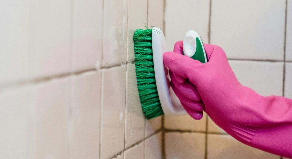 A hand in a pink glove scrubs light-colored tiled walls with a green cleaning