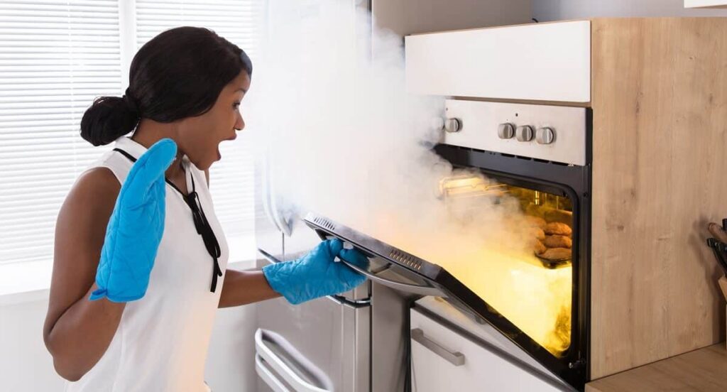A surprised woman opens a smoke-filled oven, revealing burnt food.