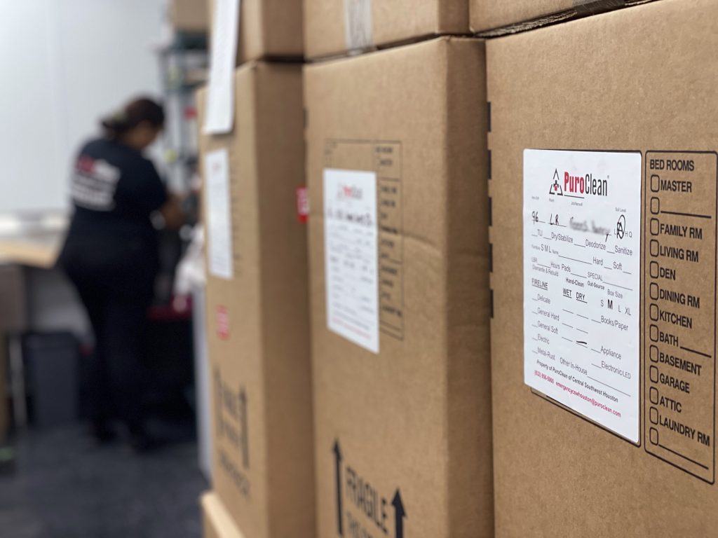 PuroClean labeled boxes are stacked in a facility, with a worker blurred in the