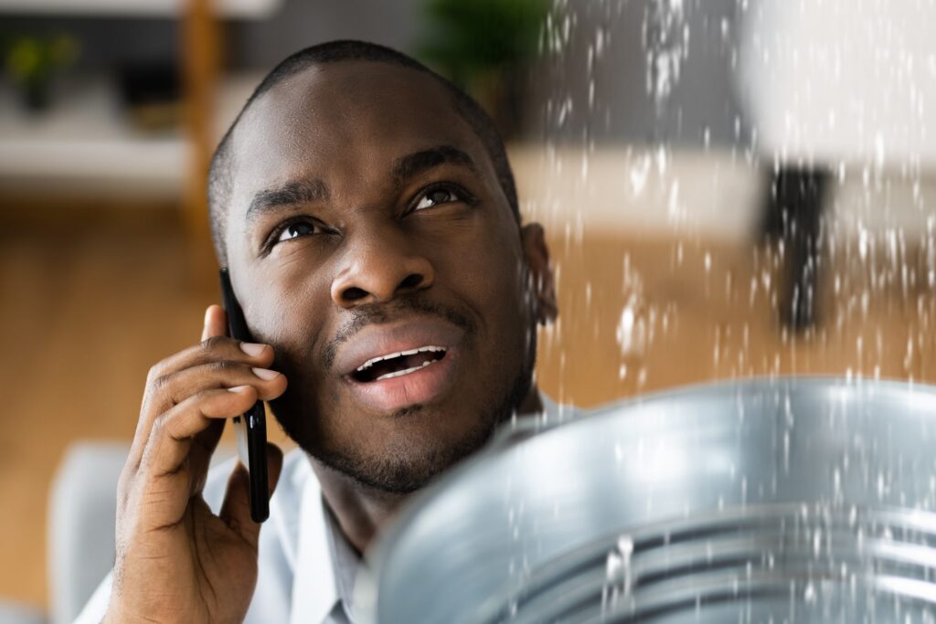 A concerned man on the phone looks up as water leaks from the ceiling into a bucket