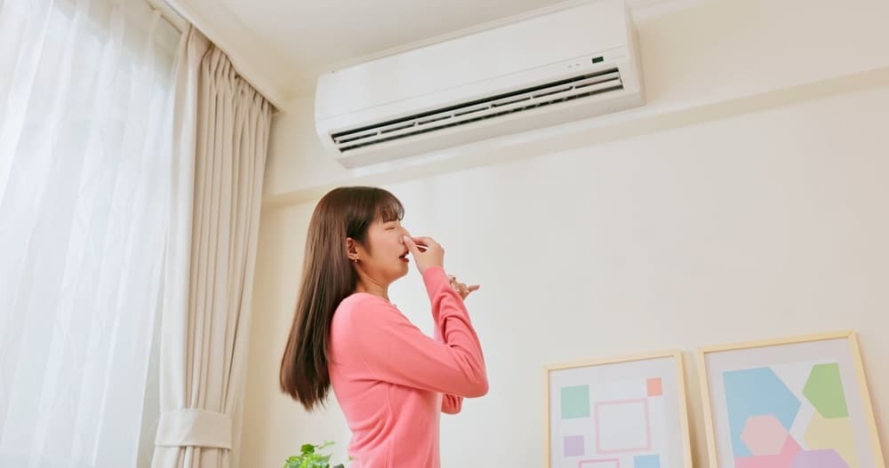 A woman pinches her nose in disgust while standing under a wall-mounted air conditioner.