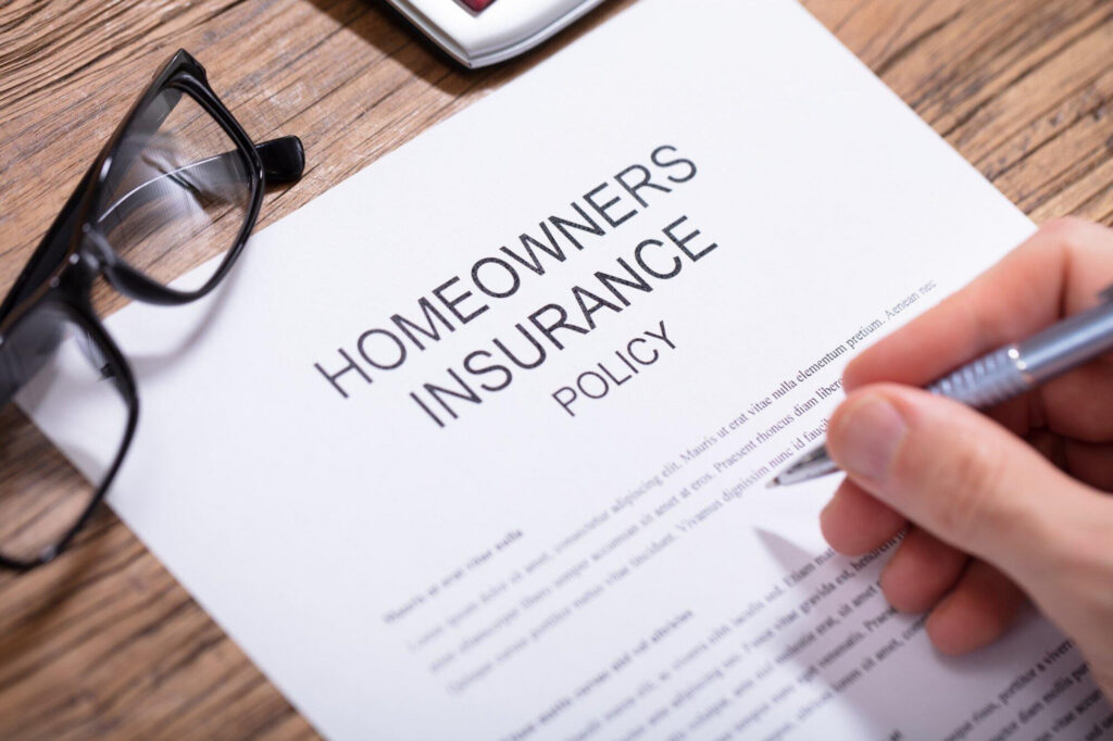 A hand holds a pen over a "HOMEOWNERS INSURANCE POLICY" document, with
