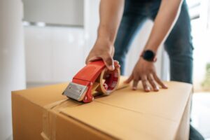 A person's hands use a red tape dispenser to seal a brown cardboard box.