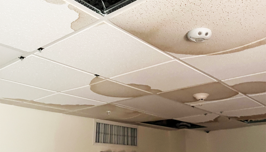 Numerous brown water stains and sagging panels cover a damaged dropped ceiling, with one tile missing.