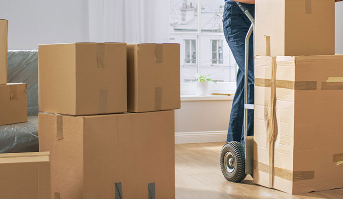 A person pushes a large cardboard box on a hand truck, surrounded by other stacked moving boxes.