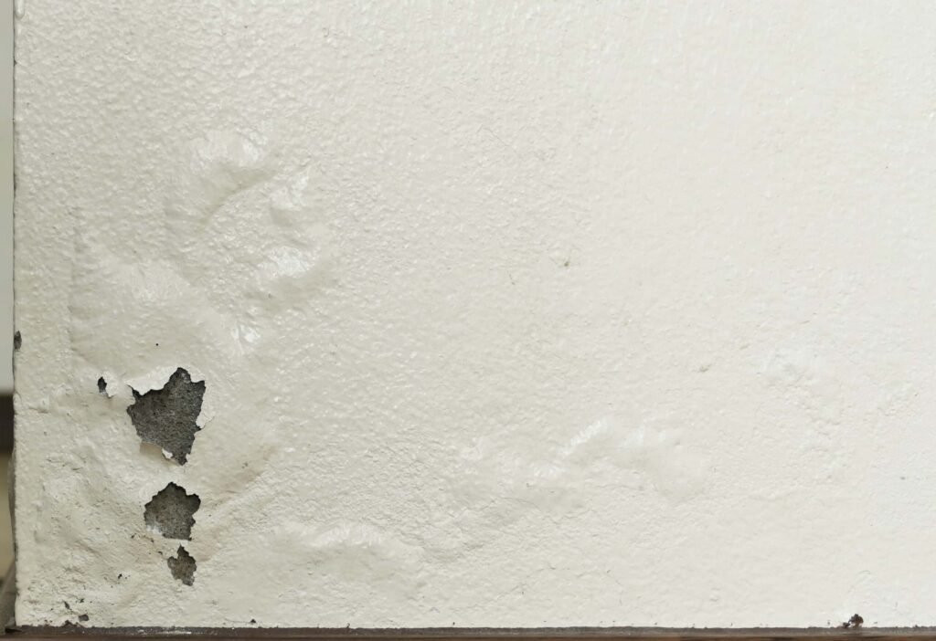 Off-white paint is peeling and bubbling on a textured wall, revealing the dark, rough