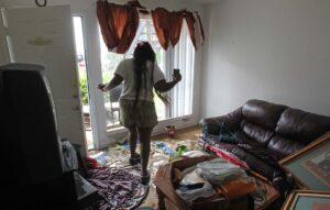 A woman surveys her flood-damaged home, standing in water amidst dislodged curtains and furniture