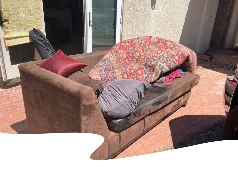 A damaged brown sofa with a red rug and pillows rests on an outdoor brick patio. TITLE
