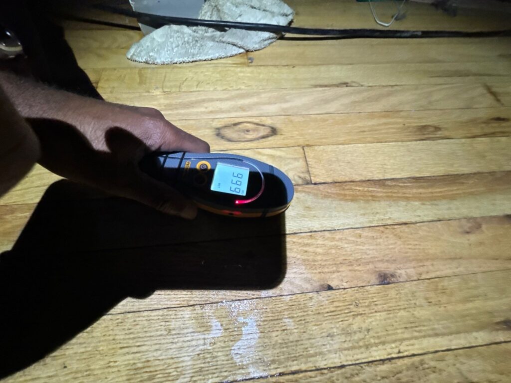 A hand holds a moisture meter displaying '99.9 WET' over a damp