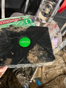 An original Xbox console and Monopoly game box are heavily caked in mud, indicating flood damage.