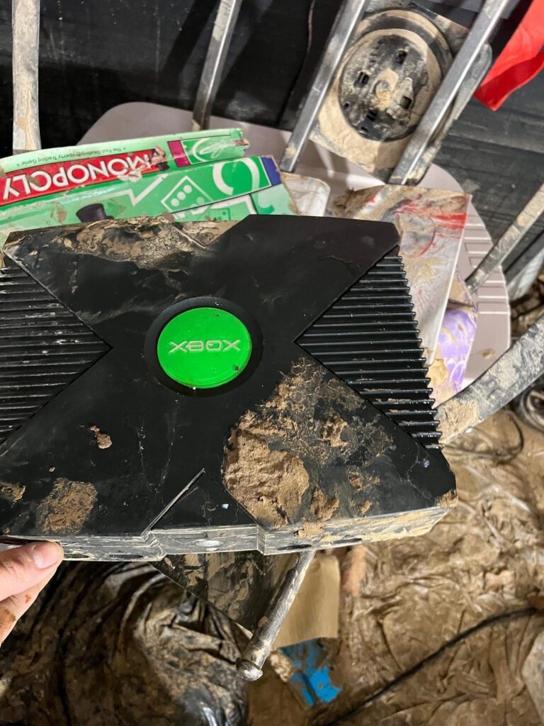 An original Xbox console and Monopoly game box are heavily caked in mud, indicating flood damage.
