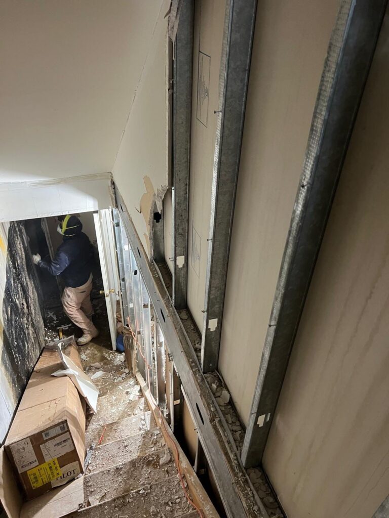 A construction worker stands in a hallway during renovation, with exposed metal wall studs and debris on