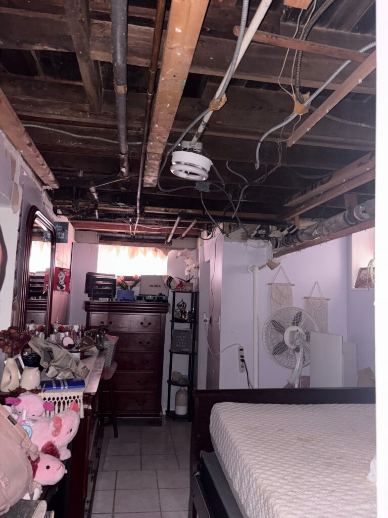 A cluttered room features an exposed ceiling with visible wooden beams, pipes, and electrical wiring