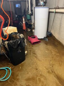 A messy basement with a wet concrete floor, a water heater, and a pink
