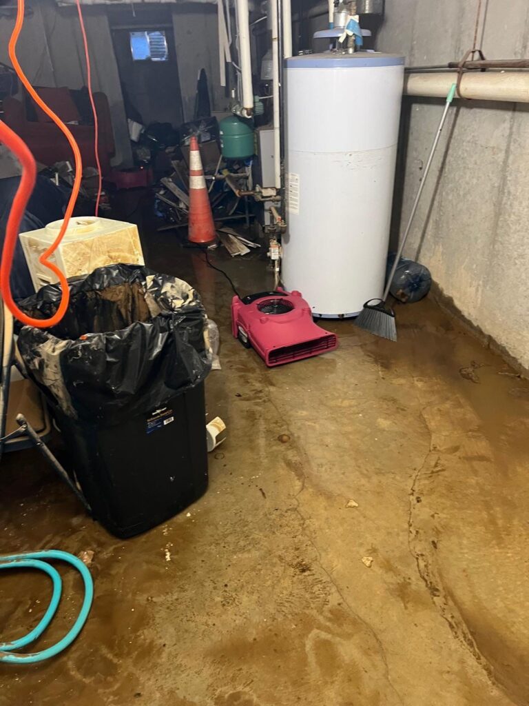 A messy basement with a wet concrete floor, a water heater, and a pink
