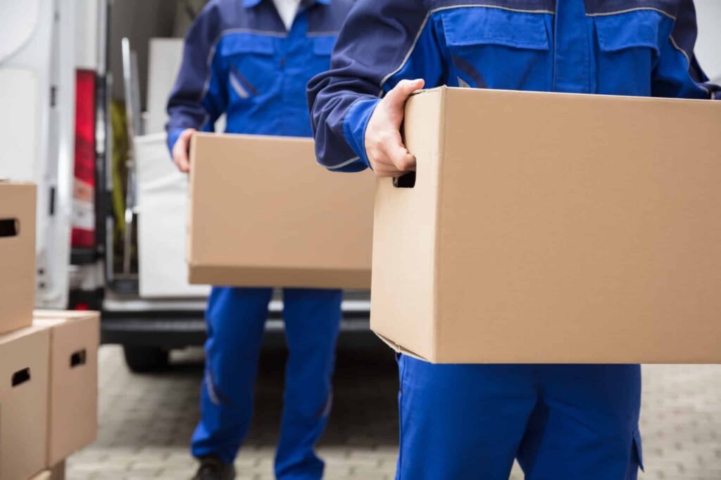 Two movers in blue uniforms carry cardboard boxes from a delivery van.
