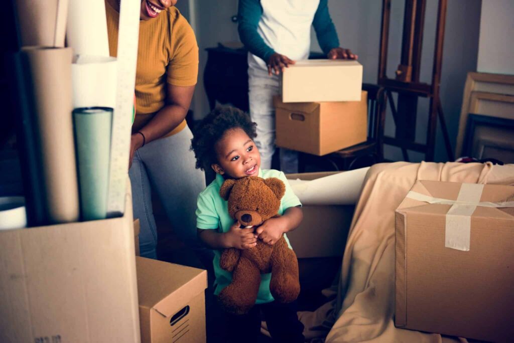 A smiling young Black child hugs a teddy bear amidst moving boxes with adults nearby. TITLE