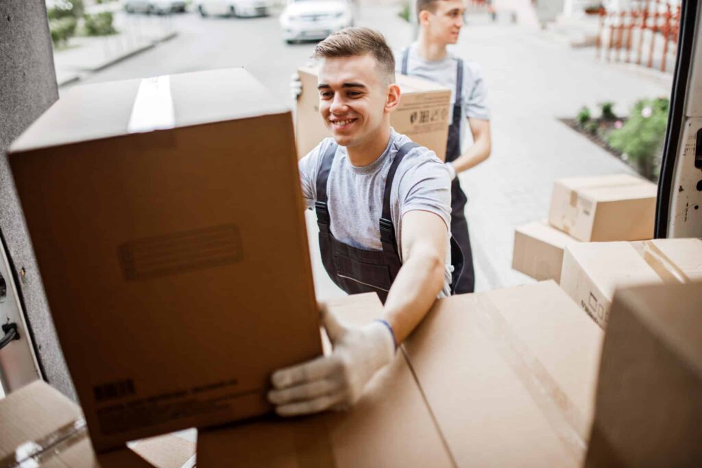 Two smiling movers load cardboard boxes into a white delivery van on a sunny day.