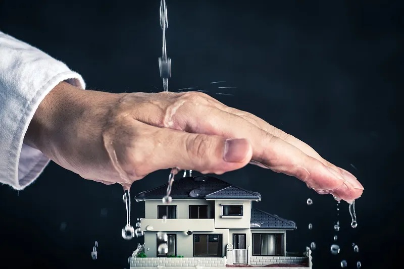A hand protects a miniature house from falling water, symbolizing home insurance. TITLE: