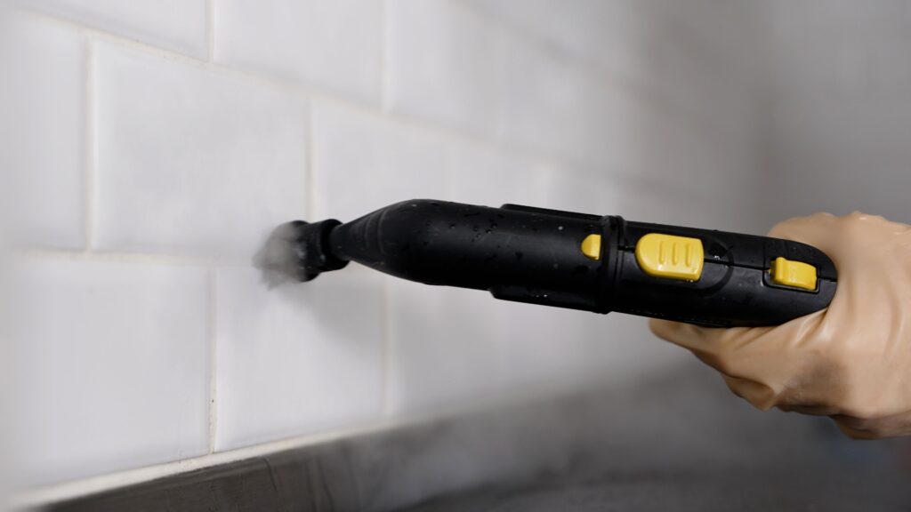 A gloved hand uses a handheld steam cleaner to clean white tiled walls.