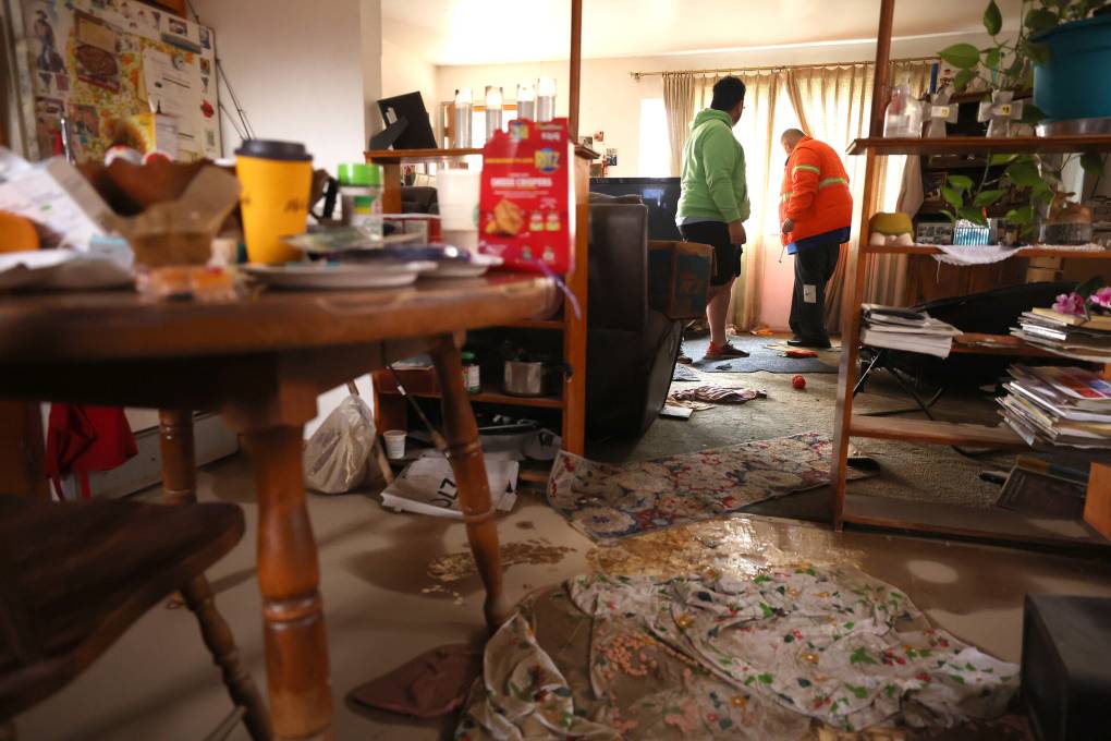 A cluttered room is flooded with water, showing soaked rugs and two men assessing the damage.
