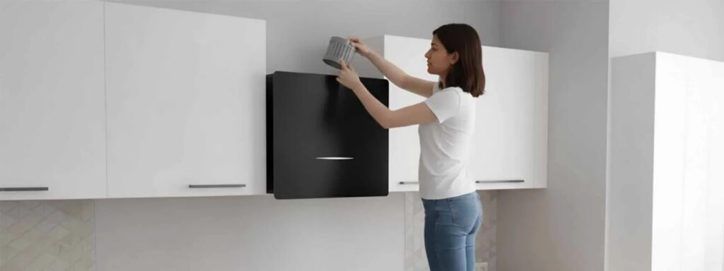 A woman installs a gray filter into a black wall-mounted kitchen air purifier. TITLE