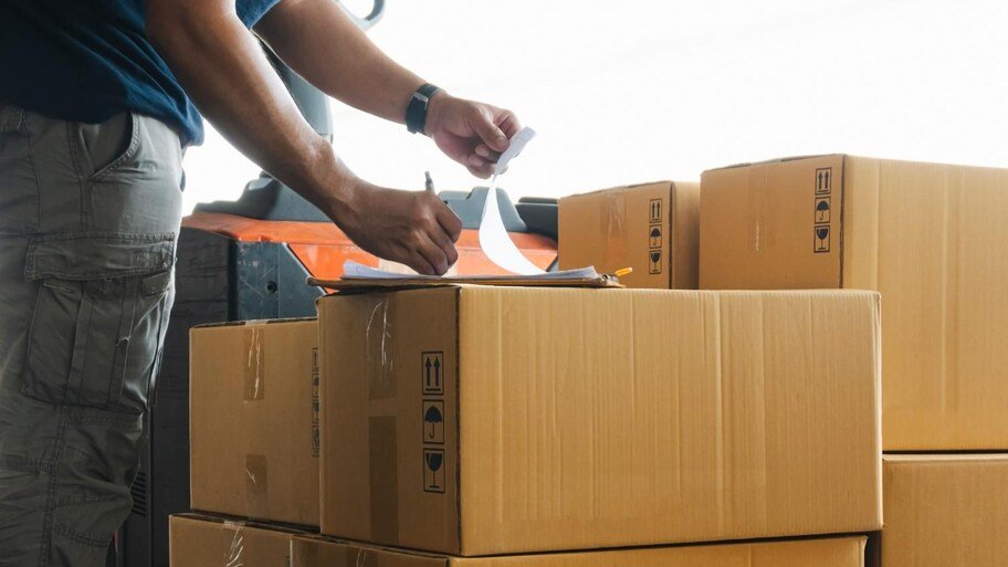 Worker signs delivery papers on a clipboard next to brown cardboard boxes.
