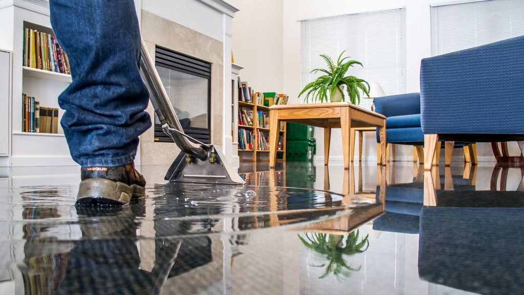 A person uses a water extractor to remove standing floodwater from a carpeted room. TITLE