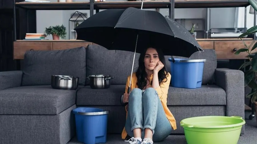 A distressed woman sits under an umbrella indoors, surrounded by pots and buckets collecting water from a