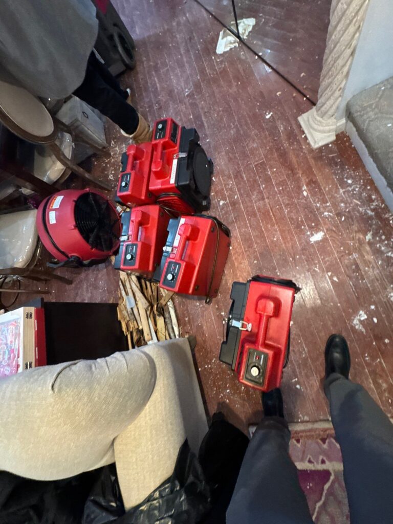 Red industrial air movers and a fan are drying a debris-strewn wooden floor. TITLE